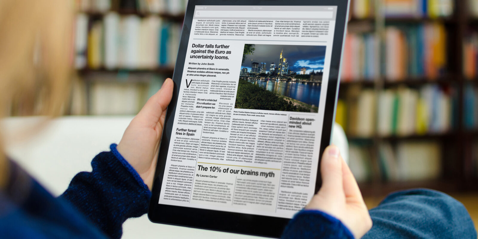 A digital newspaper on a modern tablet computer held by a young woman, who is reading the day’s news. The photo was taken by looking over the model's shoulder. A bookshelf is visible in the background of the picture. The newspaper on the device screen was created by myself in Indesign,  I own the copy right of the design and layout. The image of Frankfurt on the device is my own, I own the copy right. The text is all lorem ipsum except for the titles, which are generic and entirely fictional – these were written by myself.